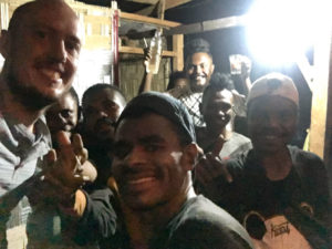 A group of boys in a dimly lit shed smiling and holding up a bottle of moonshine.