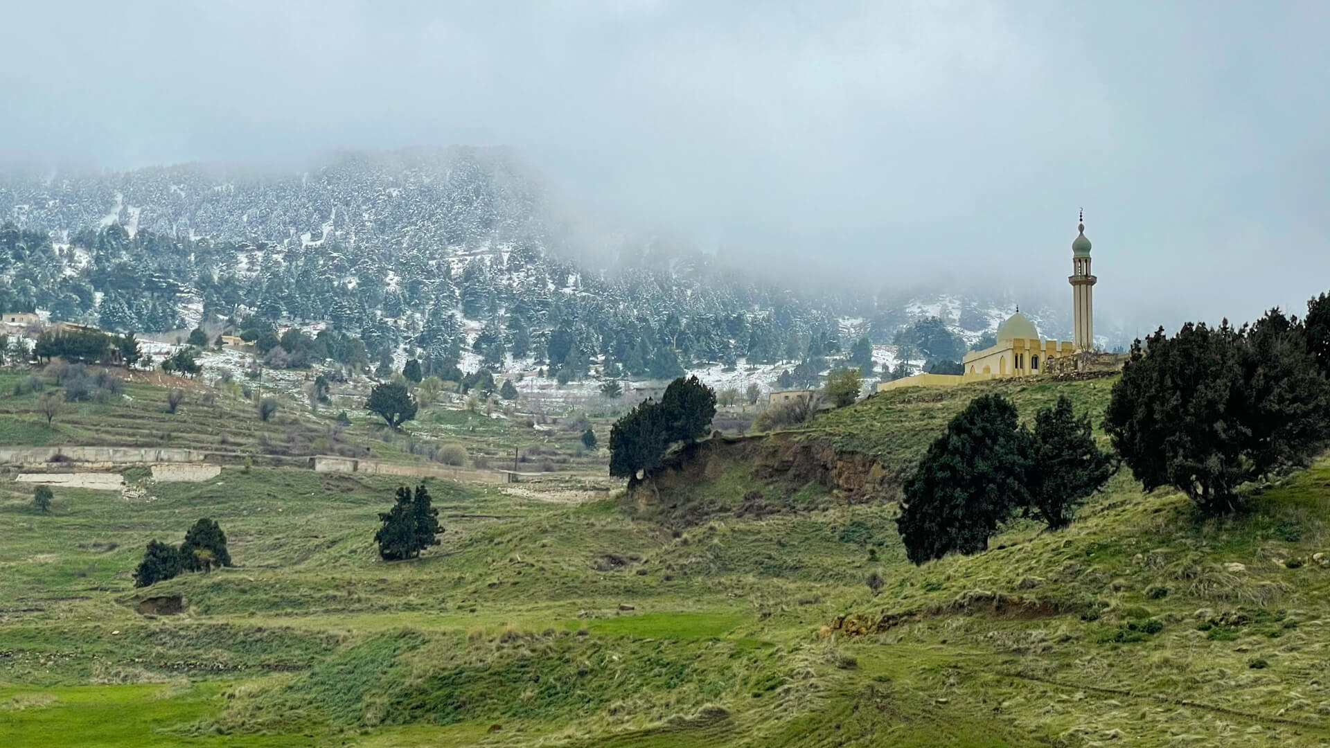 Green pastures with another mountain covered in snow rising in the distance and a mosque standing on a small hill in front.