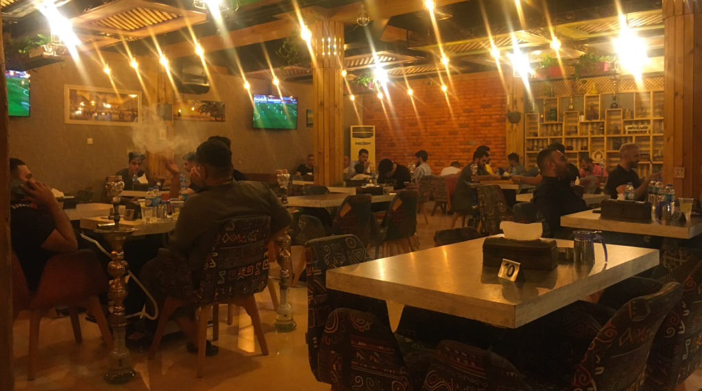 Groups of guy sit at tables smoking shisha and chatting in a dimly lit room.