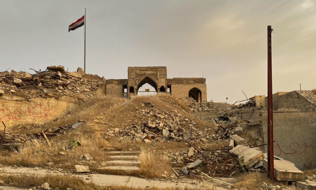 An Iraqi flag flies over the entrance to the mosque at the top of some steps.  Rubble is strewn across the surrounding area.