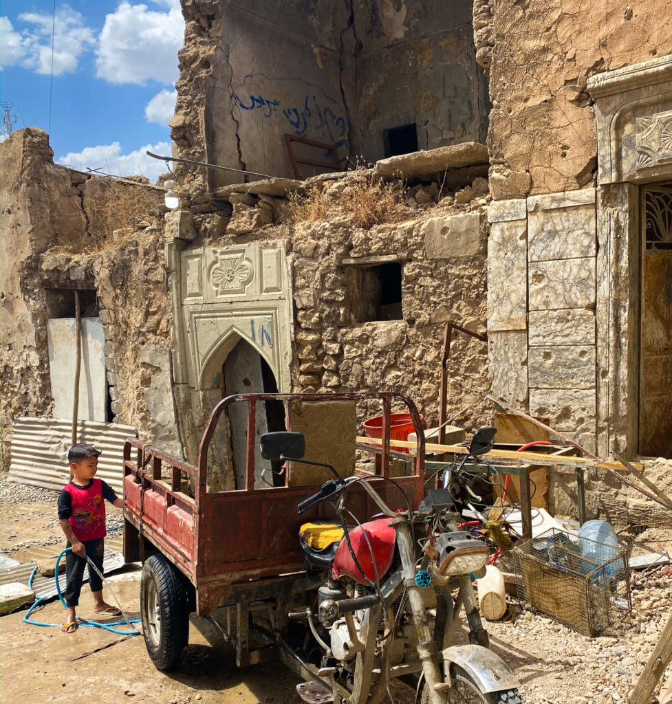 A child stands with his father's motorbike cart with ruined buildings in the background.