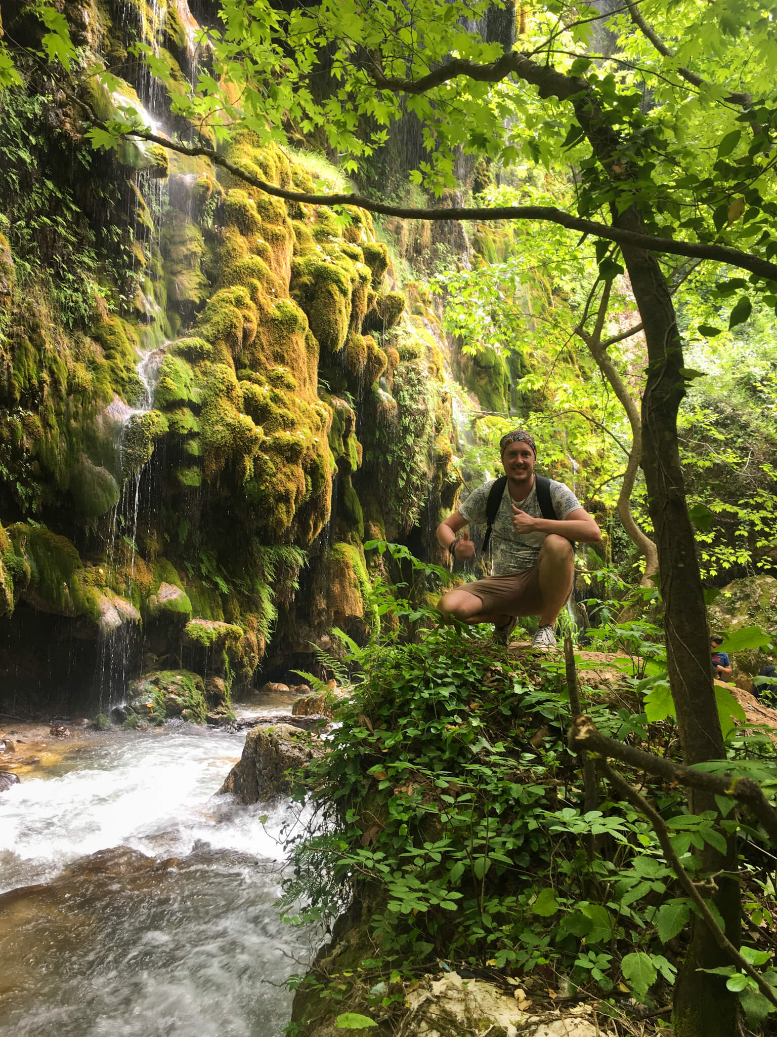 Me, squatting in a lush green forest with a small waterfall and lots of green moss in the background.