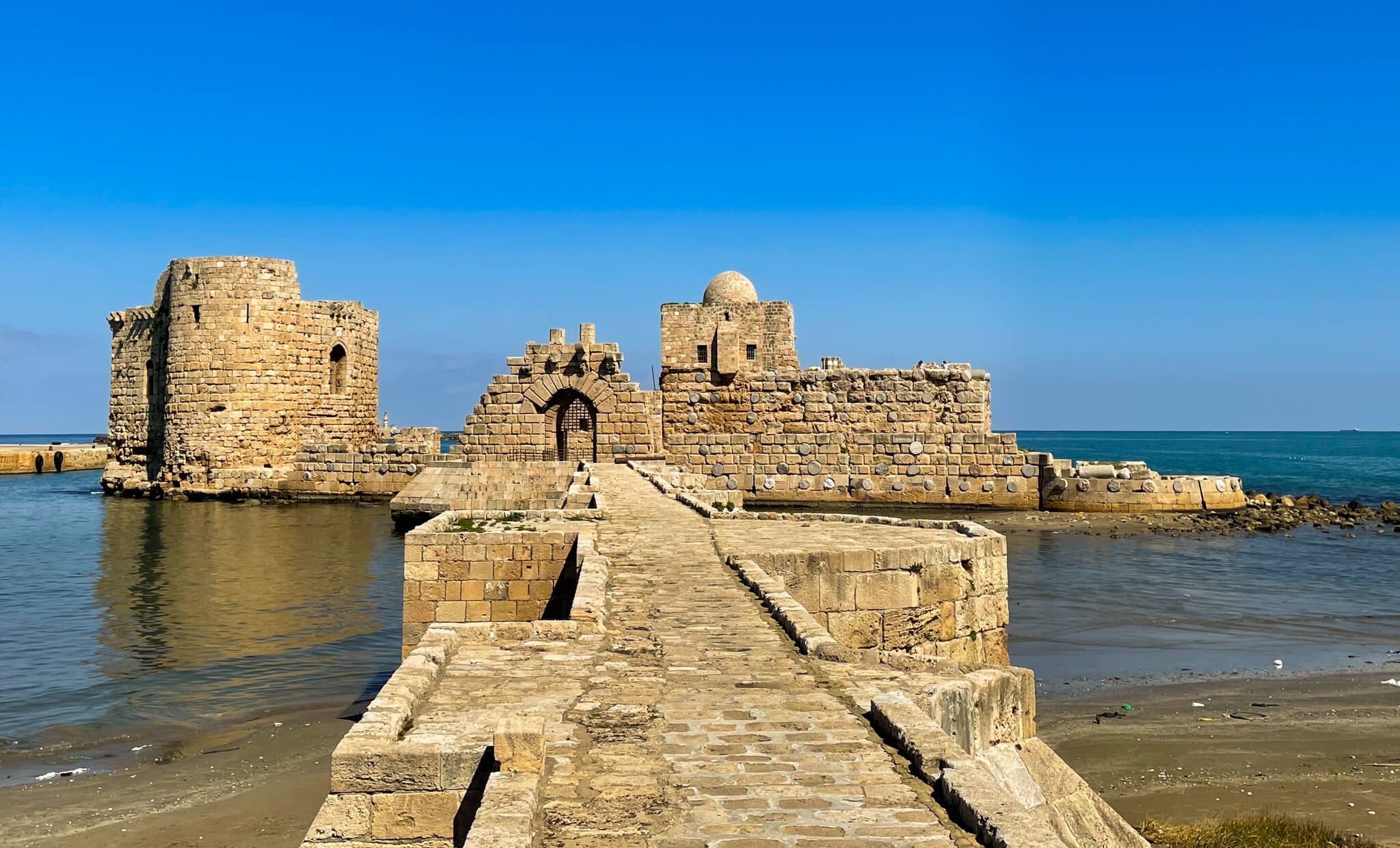 A stone castle stands in the sea with a stone walkway leading to it.