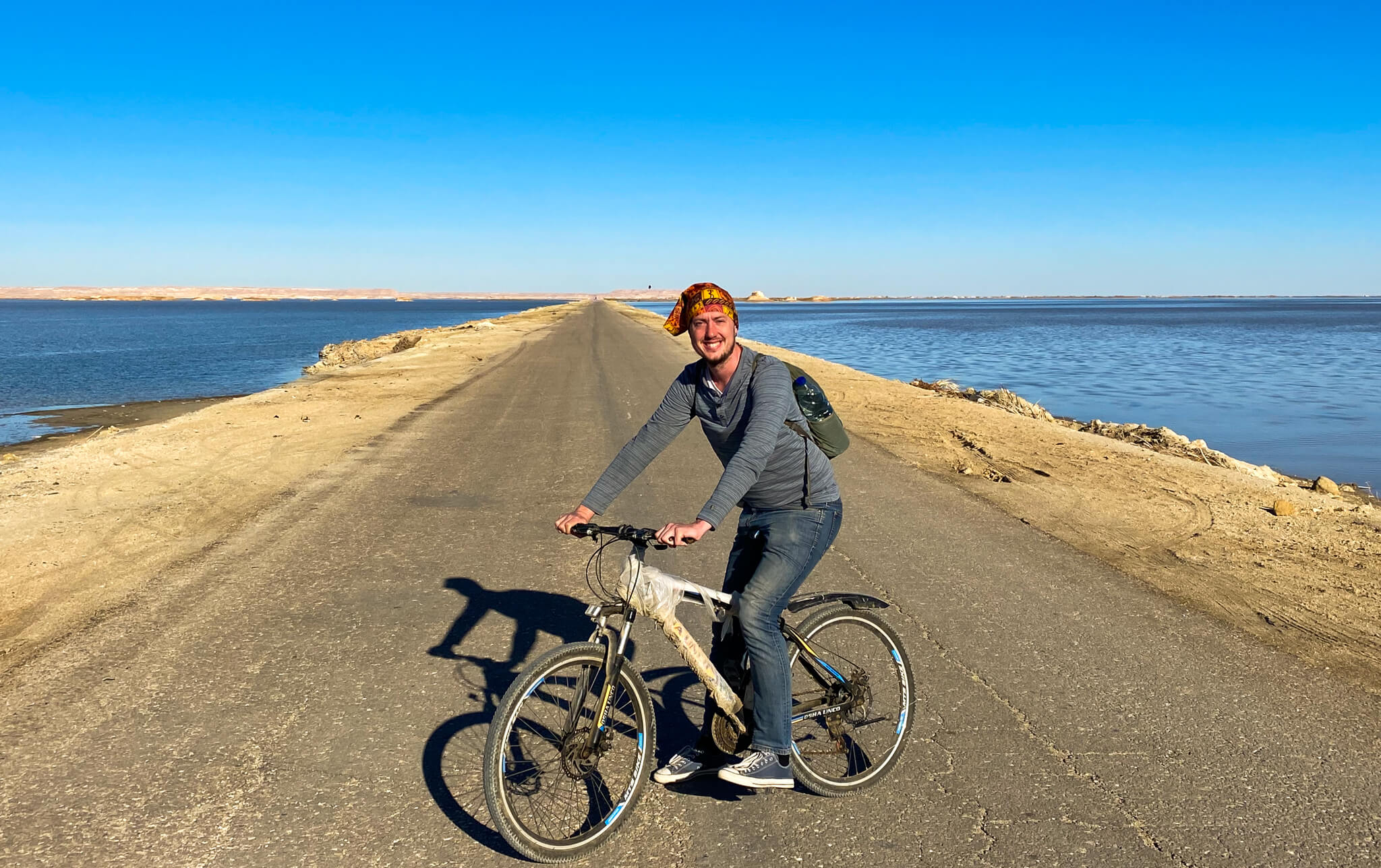 Me on a bicycle with a straight road disappearing into the distance behind me and water on either side. 