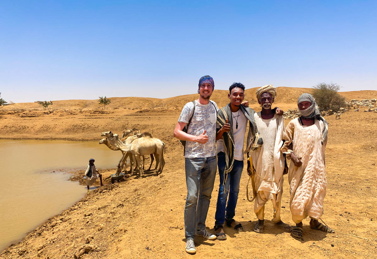 Me and three Sudanese friends in traditional costumes with camels drinking at an oasis in the background.