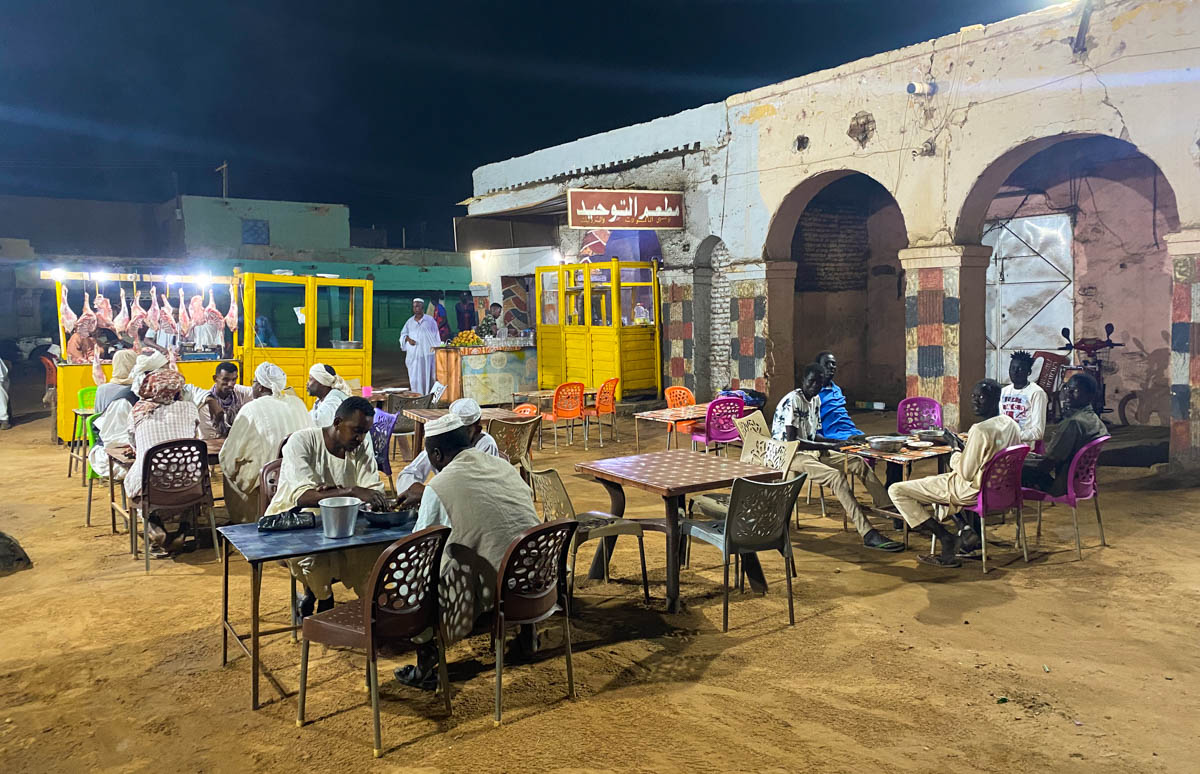 An outdoor market with pieces of meat hanging up and local people eating at tables.