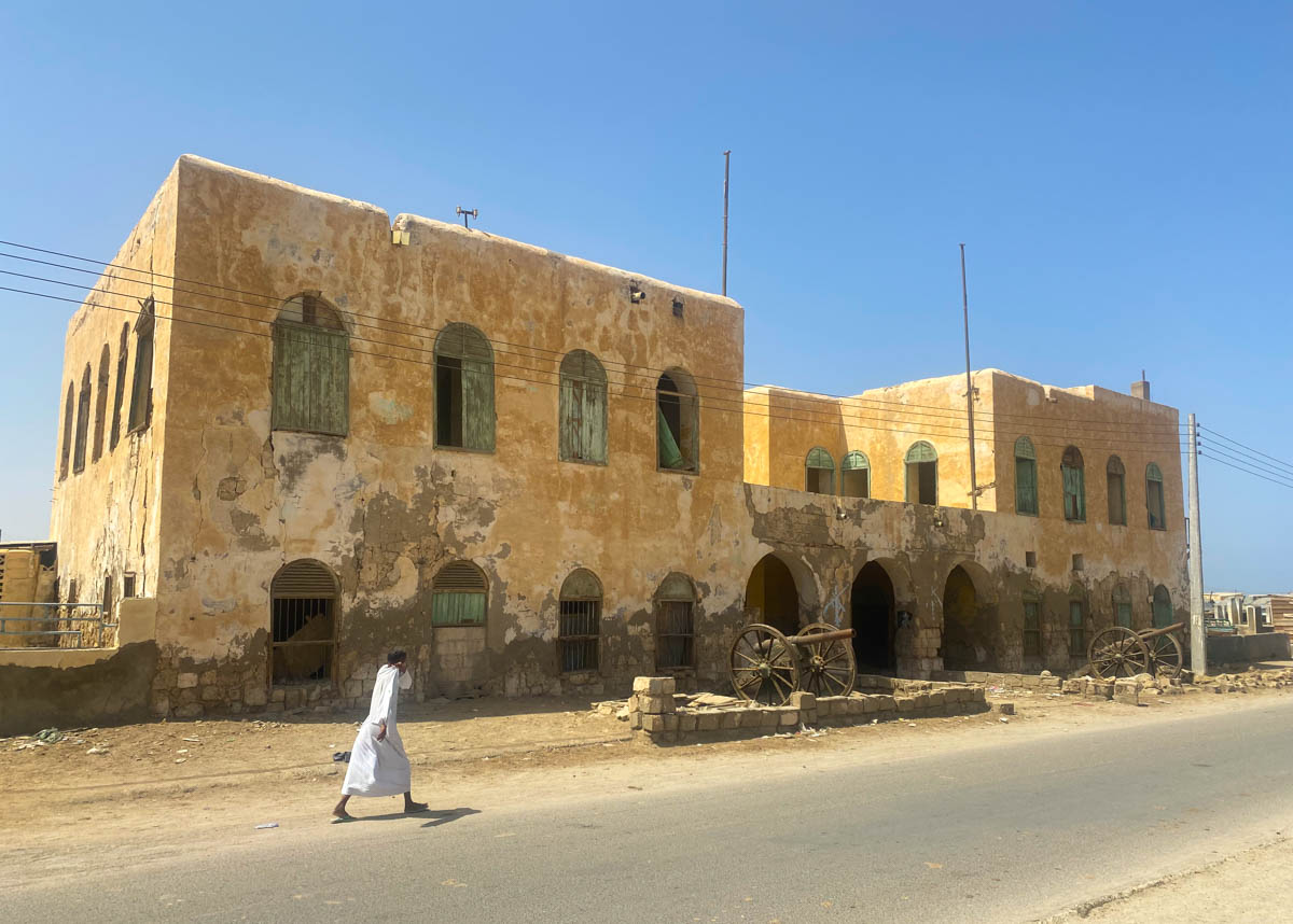 A man in local Sudanese dress walks in front of a colonial building with cannons out front